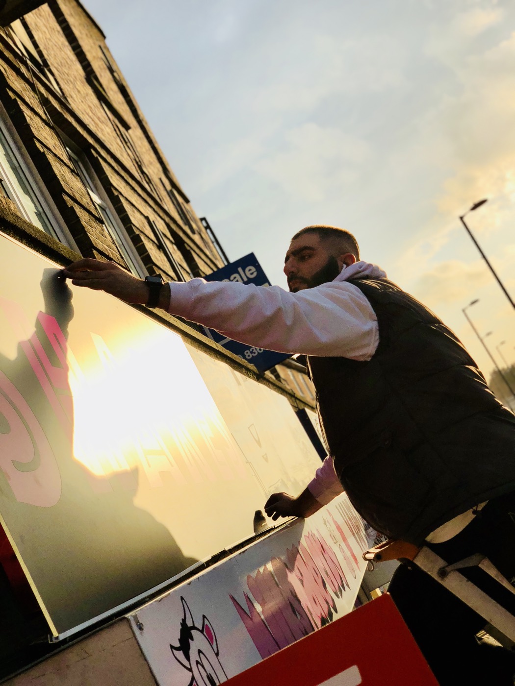 Silhouetted against a low sun, a man lifts a section of shop signage away from the fascia