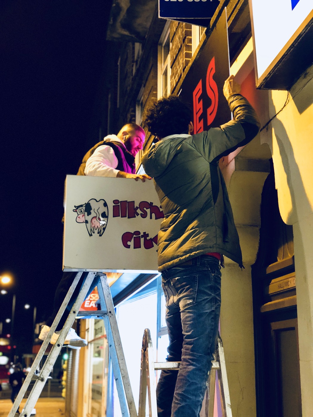 At night, a man on a ladder lowers the painted cow sign from above the shopfront